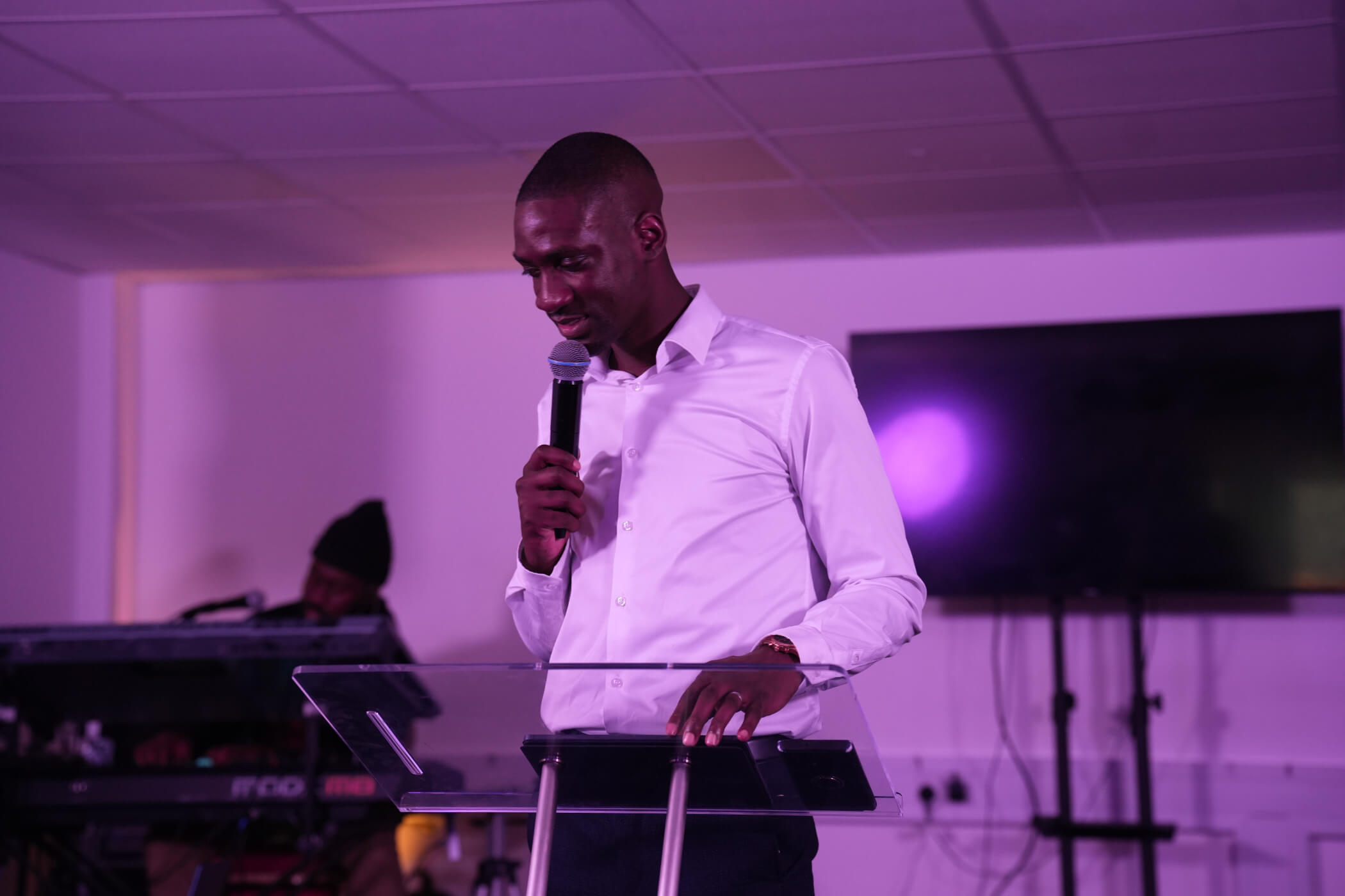 A minister preaching from a clear lectern under purple stage light
